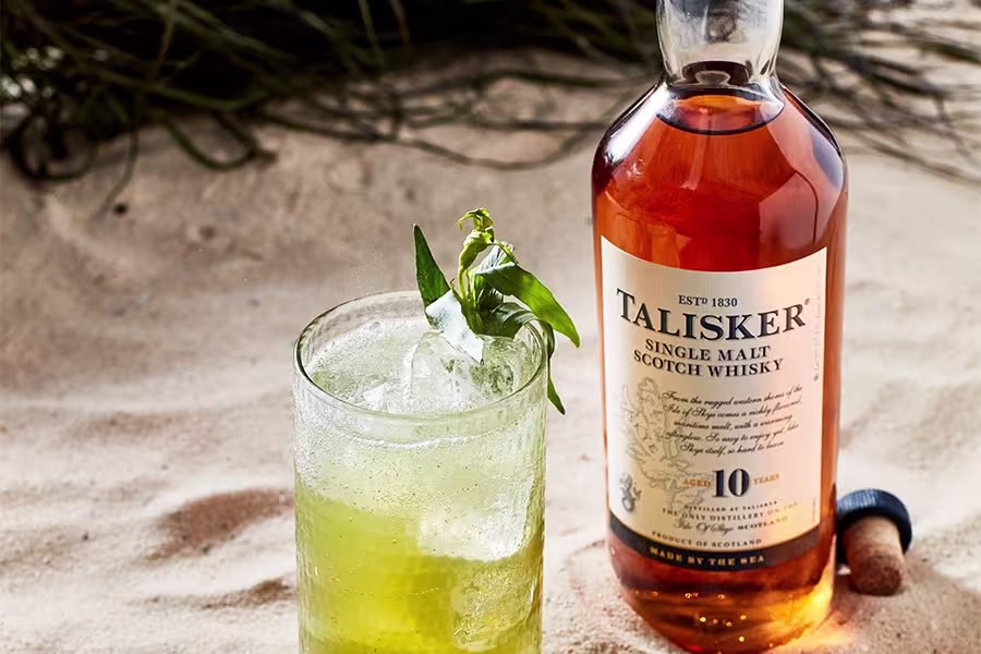 A bottle of Talisker 10-Year-Old Single Malt Scotch Whisky stands on sand next to a glass of green cocktail with ice and a leafy garnish.