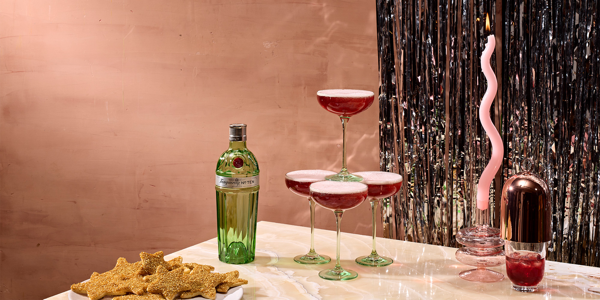 A bottle of gin, four red cocktails in coupe glasses, a plate of star-shaped cookies, and a candle in a pink holder sit on a marble surface with metallic streamers in the background.