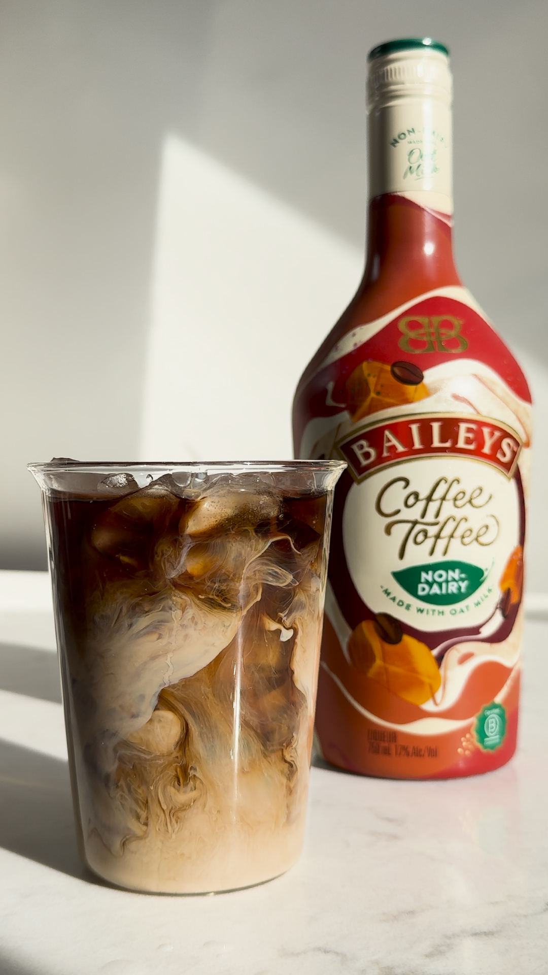 A plastic cup of iced coffee with milk swirling next to a bottle of Baileys Coffee Toffee Non-Dairy made with oat milk.