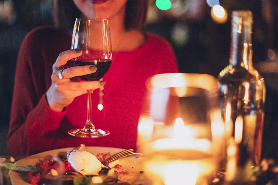 A person in a red top holds a glass of red wine at a candlelit table with a partially eaten meal and a wine bottle.