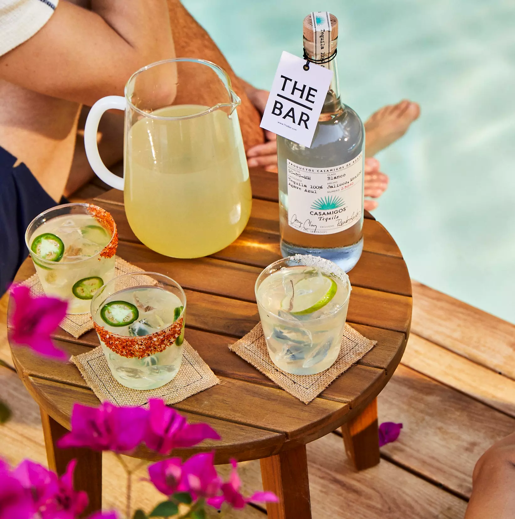 A wooden table by a pool holds a pitcher, a bottle of tequila labeled The Bar, and three cocktails with lime and chili garnish; two people sit nearby.