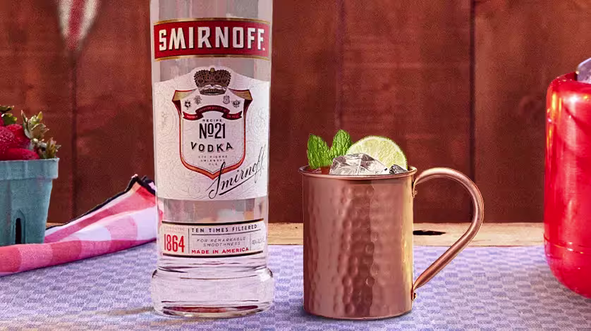 A bottle of Smirnoff No. 21 vodka stands next to a copper mug with ice, lime, and mint garnish on a checkered tablecloth.