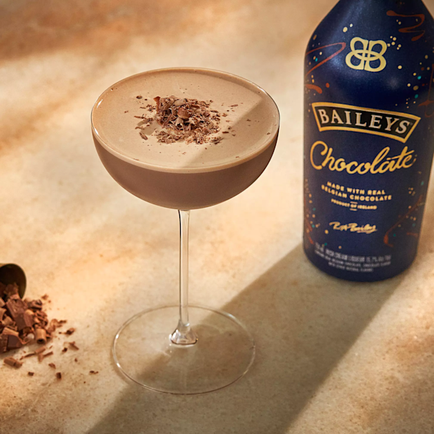 A chocolate cocktail in a coupe glass garnished with chocolate shavings, next to a bottle of Baileys Chocolate liqueur on a beige surface.