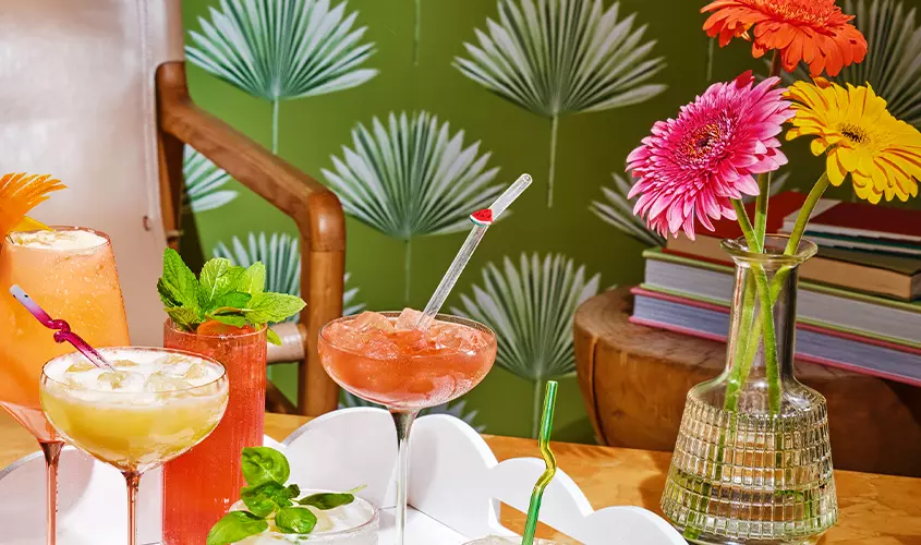 A selection of colorful cocktails in various glasses sits on a white tray next to a vase of gerbera daisies and a stack of books against a green palm-leaf wallpaper background.