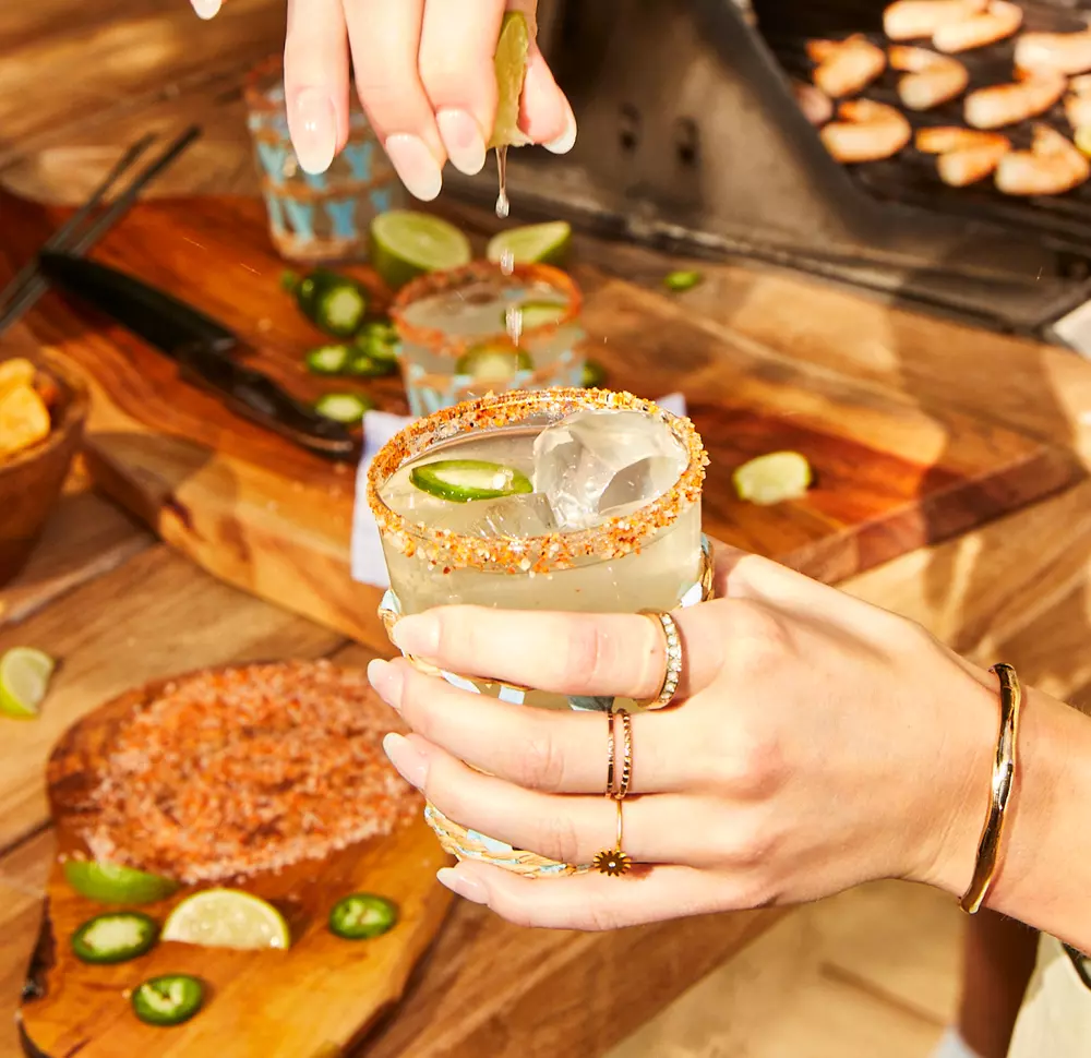 A person squeezes a lime over an iced cocktail in a glass with a spiced rim, surrounded by sliced jalapeños, lime wedges, and food on a wooden table.