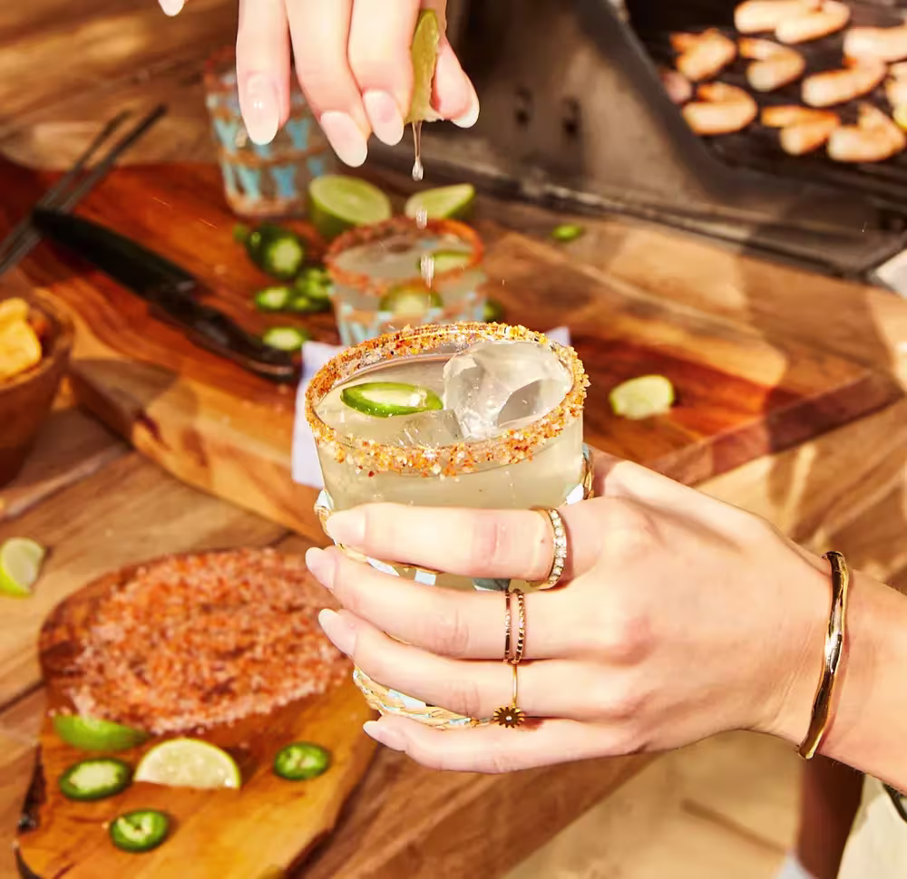 A person squeezes a lime over an iced cocktail in a glass with a spiced rim, surrounded by sliced jalapeños, lime wedges, and food on a wooden table.