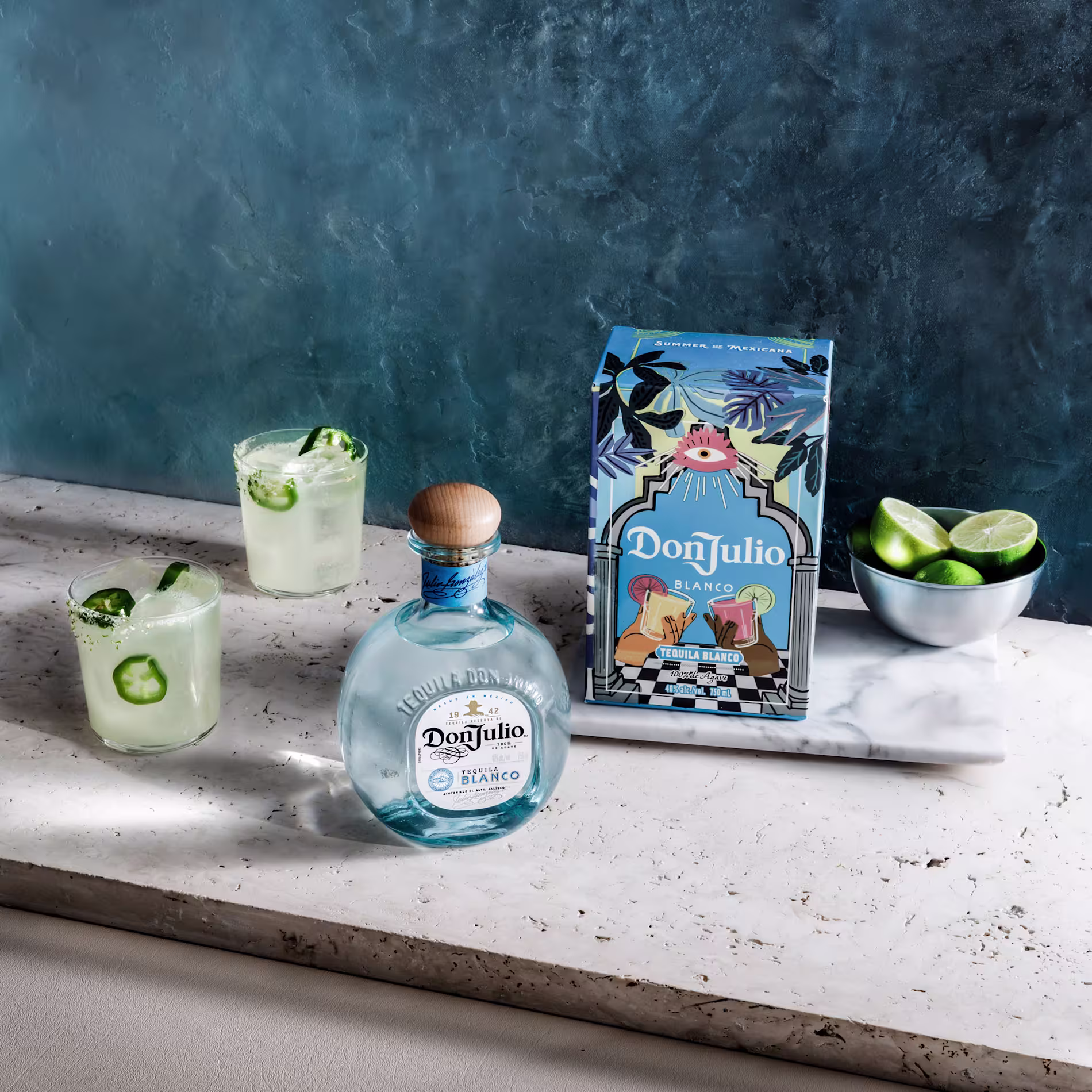 A bottle of Don Julio Blanco tequila, its decorative box, two cocktails with lime and cucumber, and a bowl of lime wedges on a marble surface against a blue background.