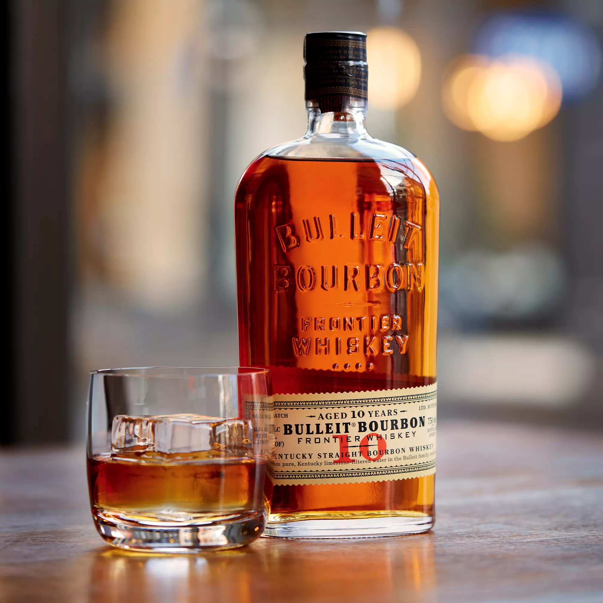 A bottle of Bulleit Bourbon Frontier Whiskey and a glass of bourbon with ice are placed on a wooden surface.