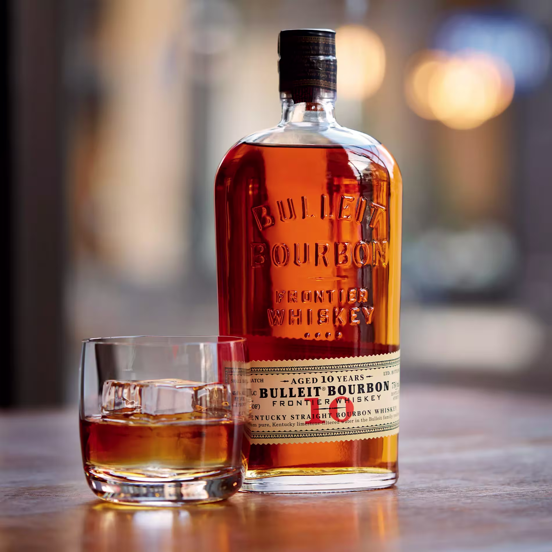 A bottle of Bulleit Bourbon Frontier Whiskey and a glass of bourbon with ice are placed on a wooden surface.