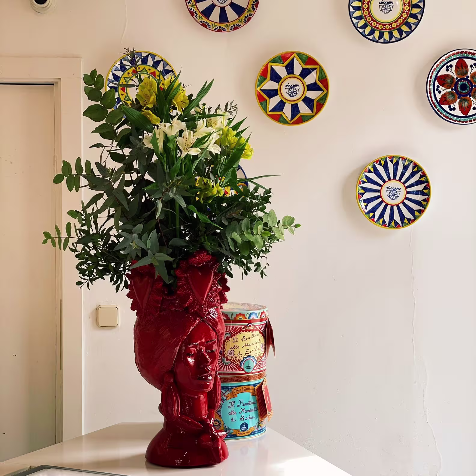 Interior de Zùccaru con pared decorada con platos sicilianos, jarrón rojo con forma de cabeza femenina y flores frescas junto a coloridas latas artesanales.
