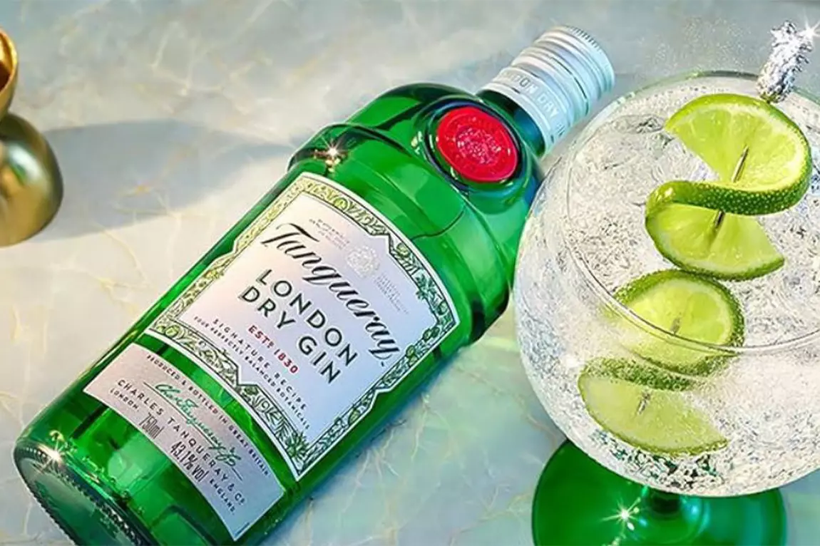 A bottle of Tanqueray London Dry Gin lies next to a glass of gin and tonic garnished with lime slices and a lime twist.