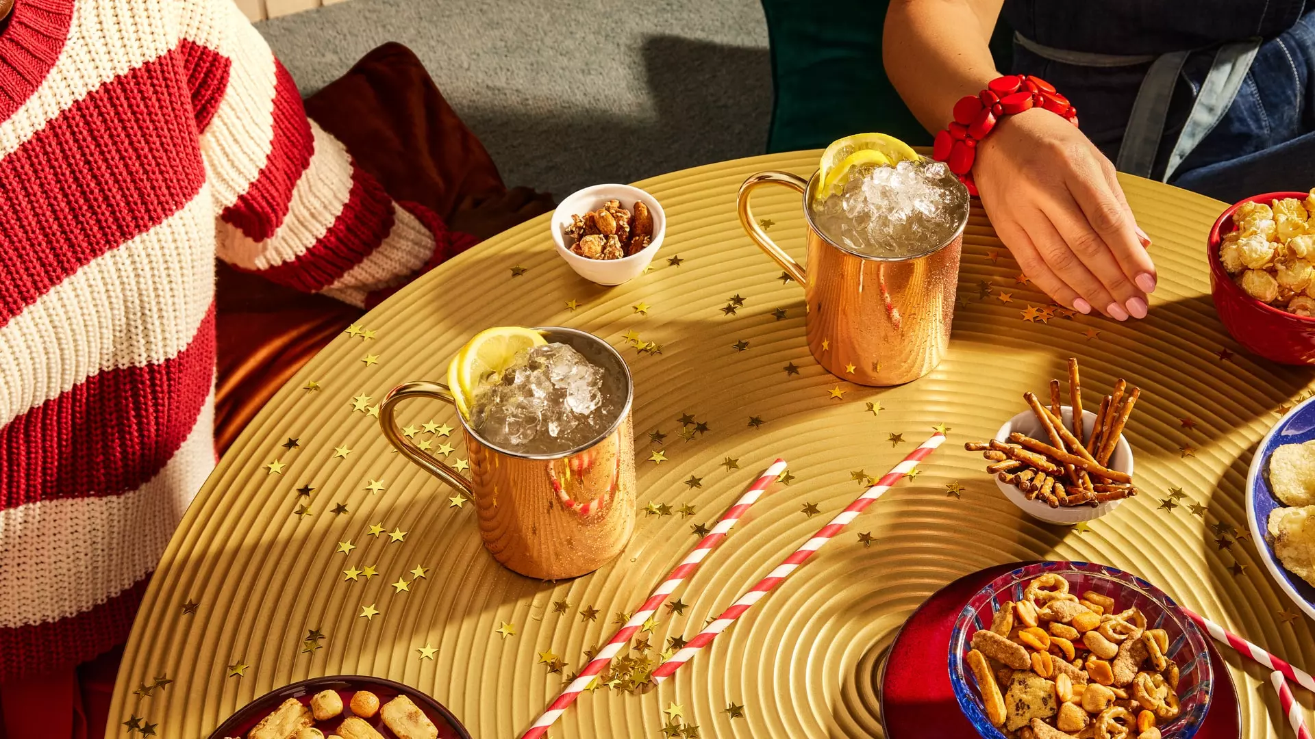 Two people sit at a gold table with two Moscow mule mugs, snacks in bowls, paper straws, and gold star confetti scattered across the surface.