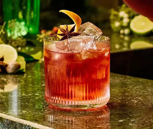 A glass of red cocktail with ice, garnished with an orange peel and star anise, sits on a stone countertop with lime slices and greenery in the background.