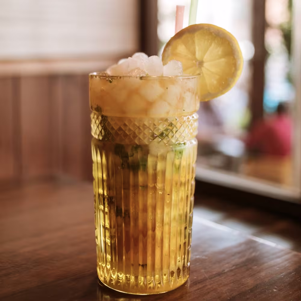 La imagen muestra un vaso de lo que parece un mojito, adornado con una rodaja de limón y una pajilla, colocado sobre una superficie de madera.