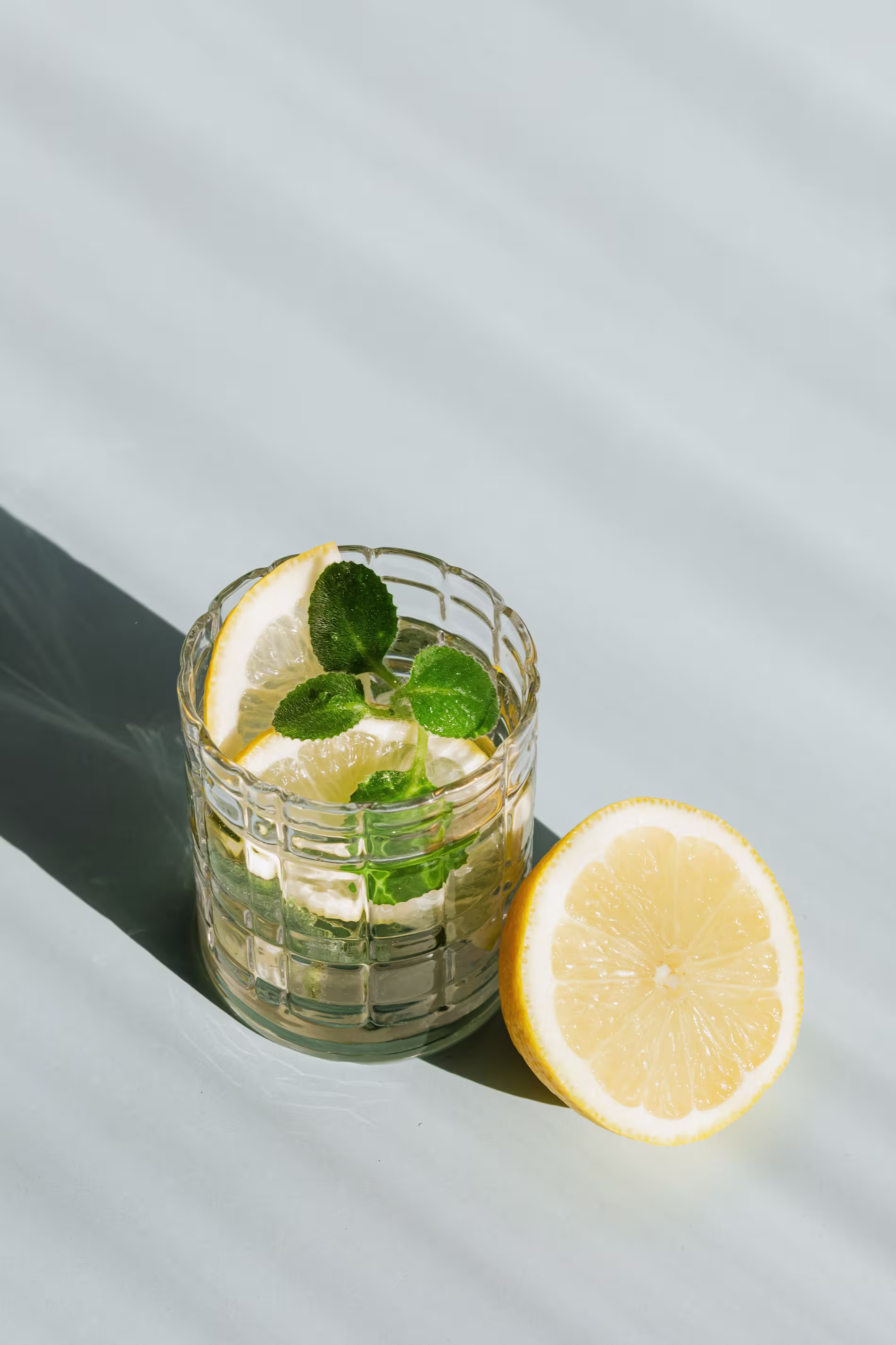 A glass of water with lemon slices and mint leaves beside a halved lemon on a light surface in bright sunlight.