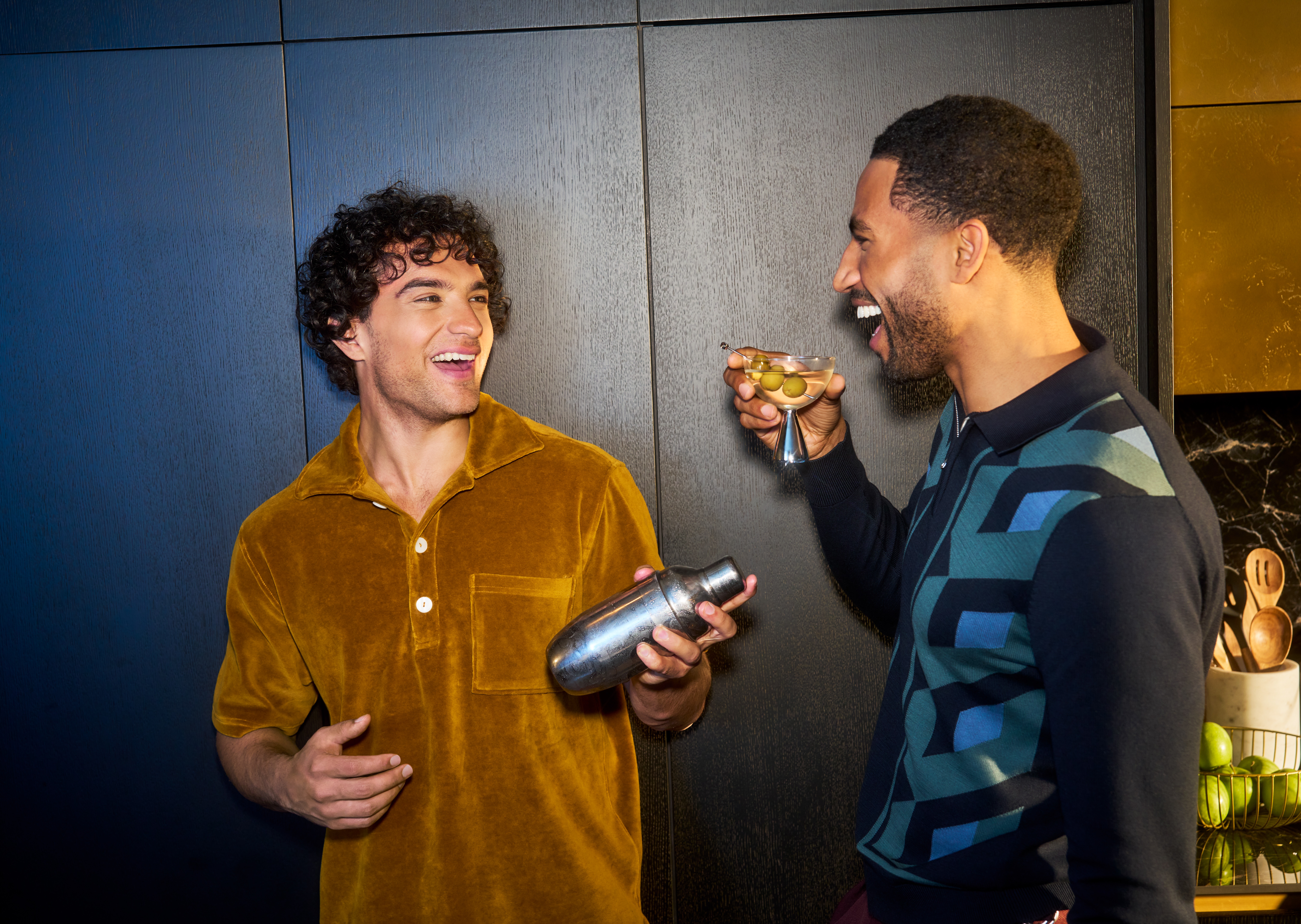 Two men enjoying cocktails, one on the left is shaking a cocktail shaker.