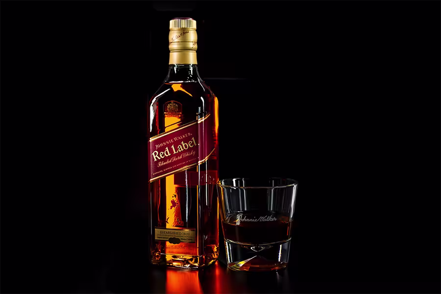 A bottle of Johnnie Walker Red Label whisky stands next to a glass containing whisky, both set against a black background.