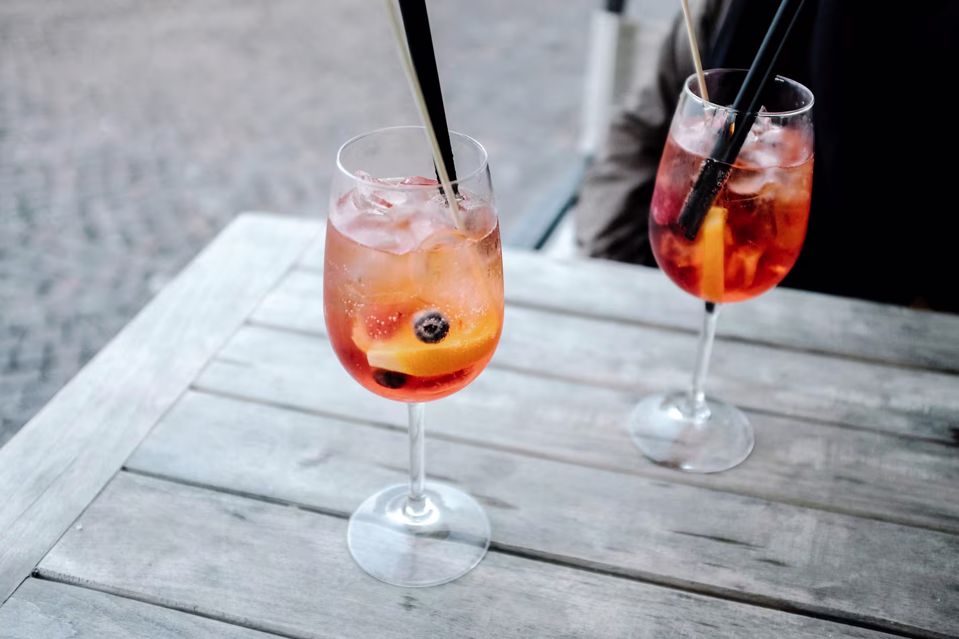 Two glasses of iced cocktails with fruit and straws on a wooden table outdoors.
