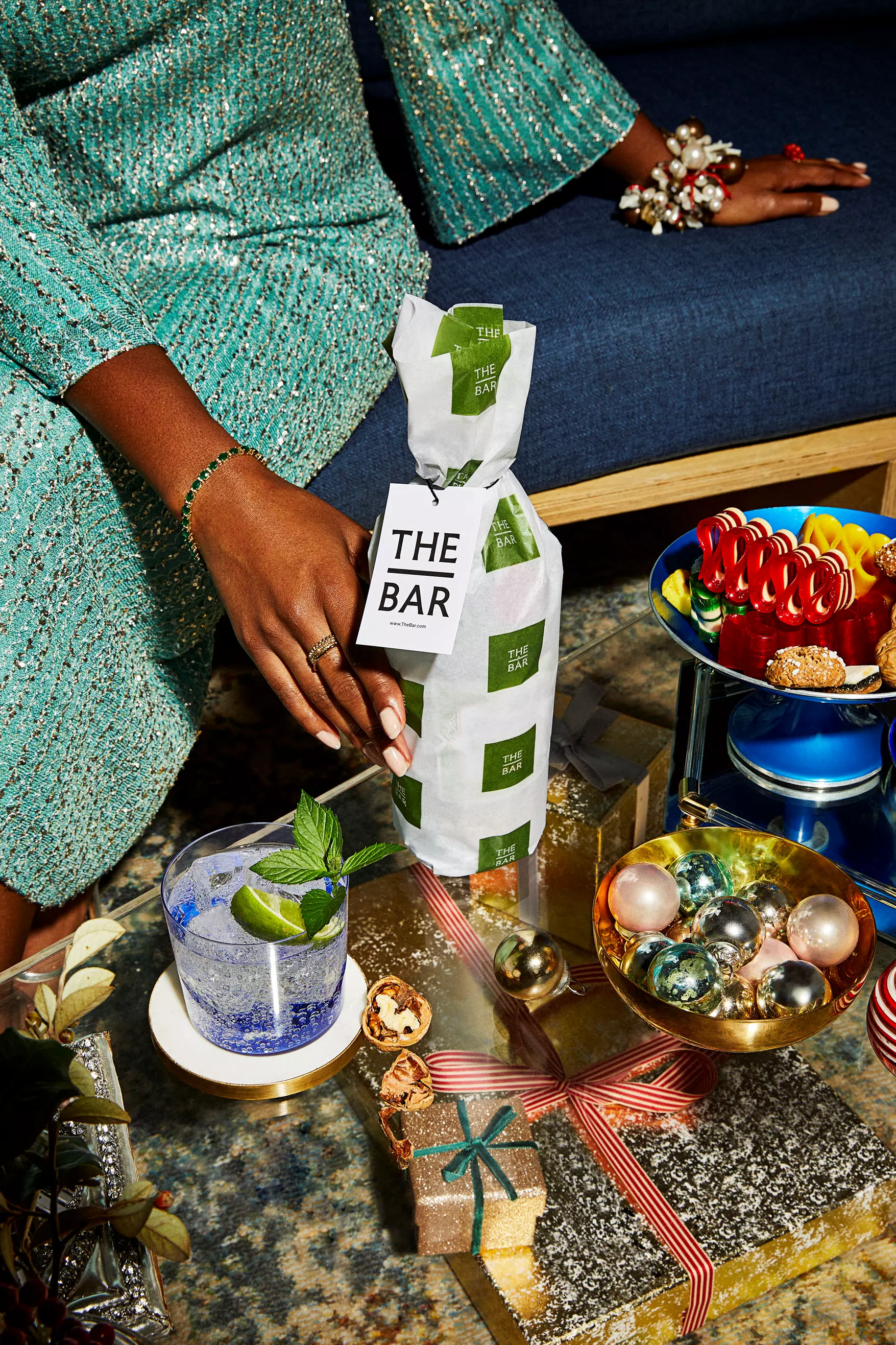 A person in a sparkly green dress holds a bottle labeled THE BAR on a table with holiday decorations, wrapped gifts, a cocktail, and festive treats.