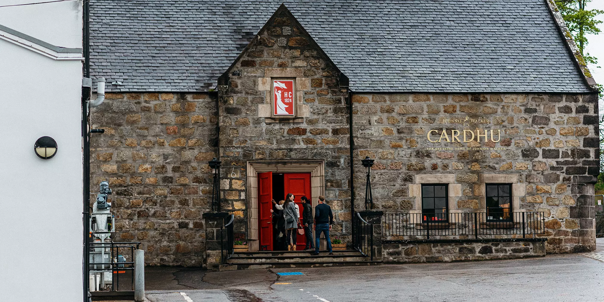 Stone building with a sign reading Cardhu, featuring a red door, a statue on the left, and several people standing at the entrance.