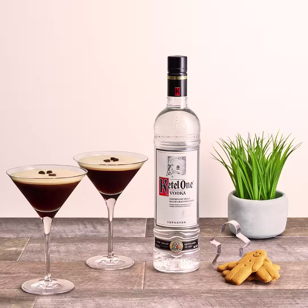 A bottle of Ketel One vodka with two espresso martinis, a small potted plant, a cookie, and a cocktail stirrer on a wooden surface.