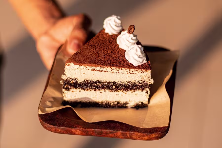 A hand holds a wooden plate with a slice of layered cake topped with whipped cream, cocoa powder, and a coffee bean.