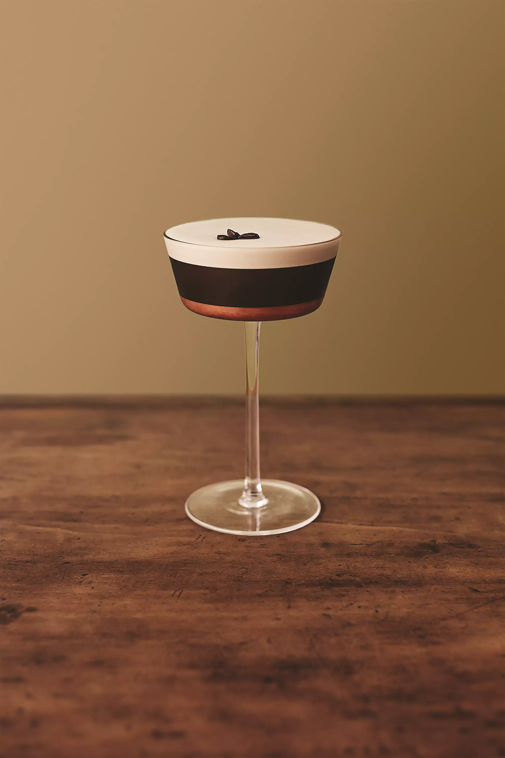A coupe glass filled with a creamy, layered cocktail garnished with three coffee beans, placed on a wooden surface against a plain beige background.
