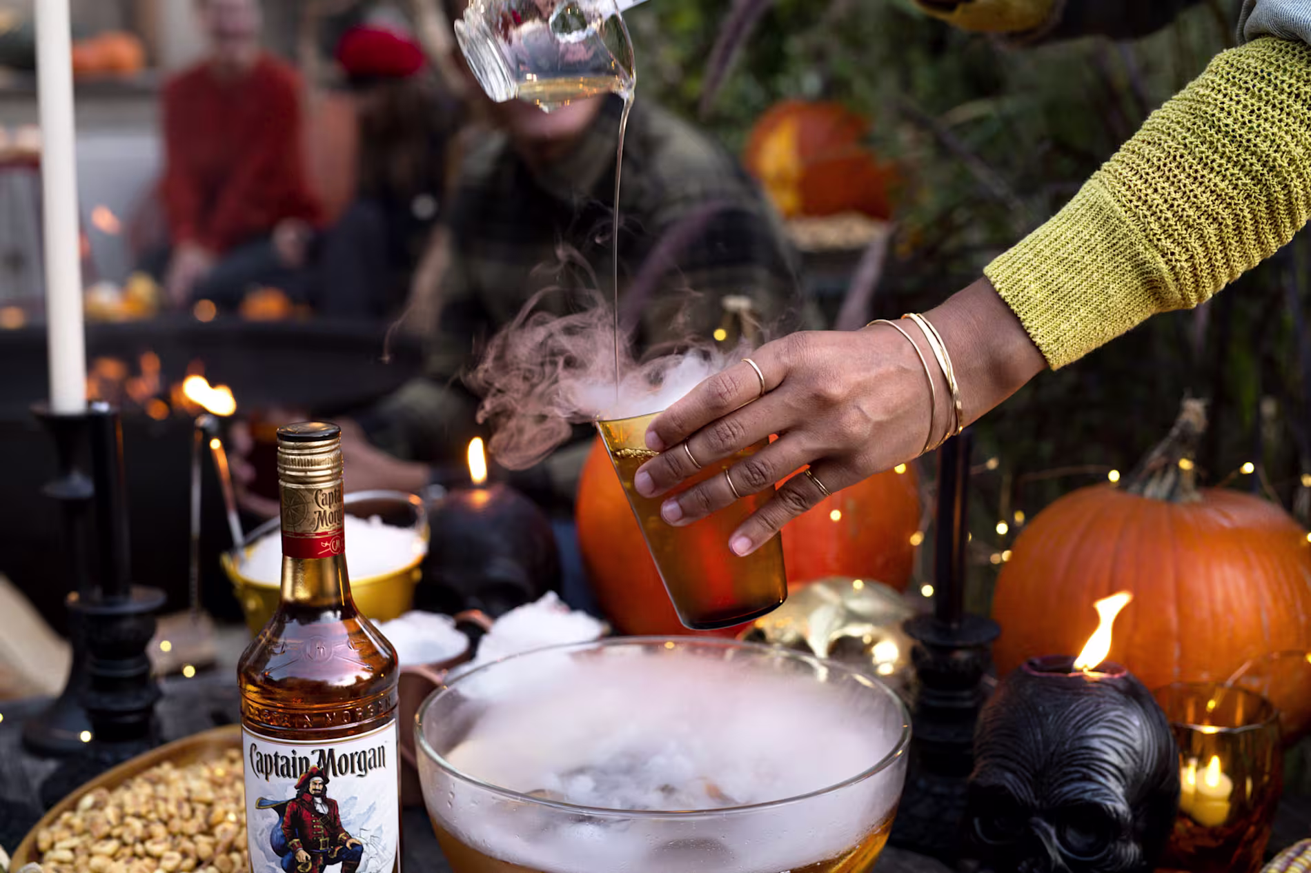A person pours liquid into a smoky drink from a bowl near a bottle of Captain Morgan, surrounded by pumpkins, candles, and Halloween decorations.