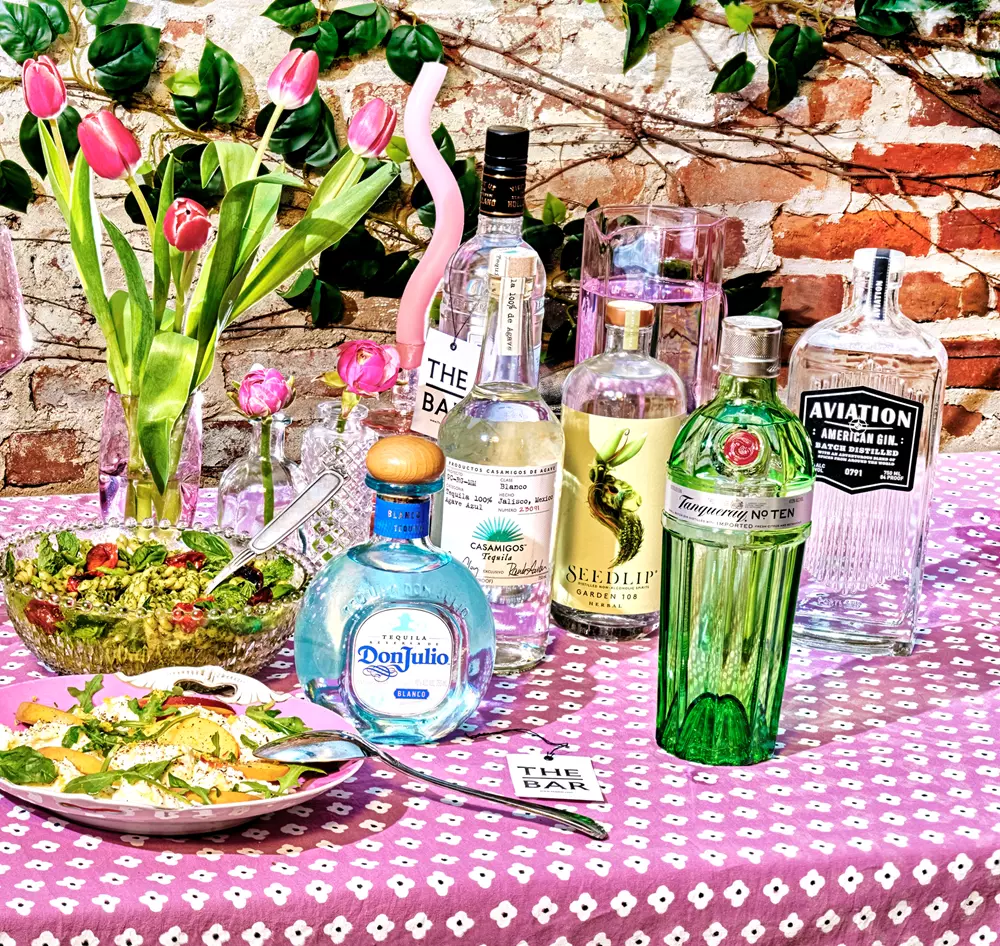 Bottles of liquor, a salad, tulip flowers, and glasses sit on a patterned tablecloth outdoors against a brick wall with green vines.