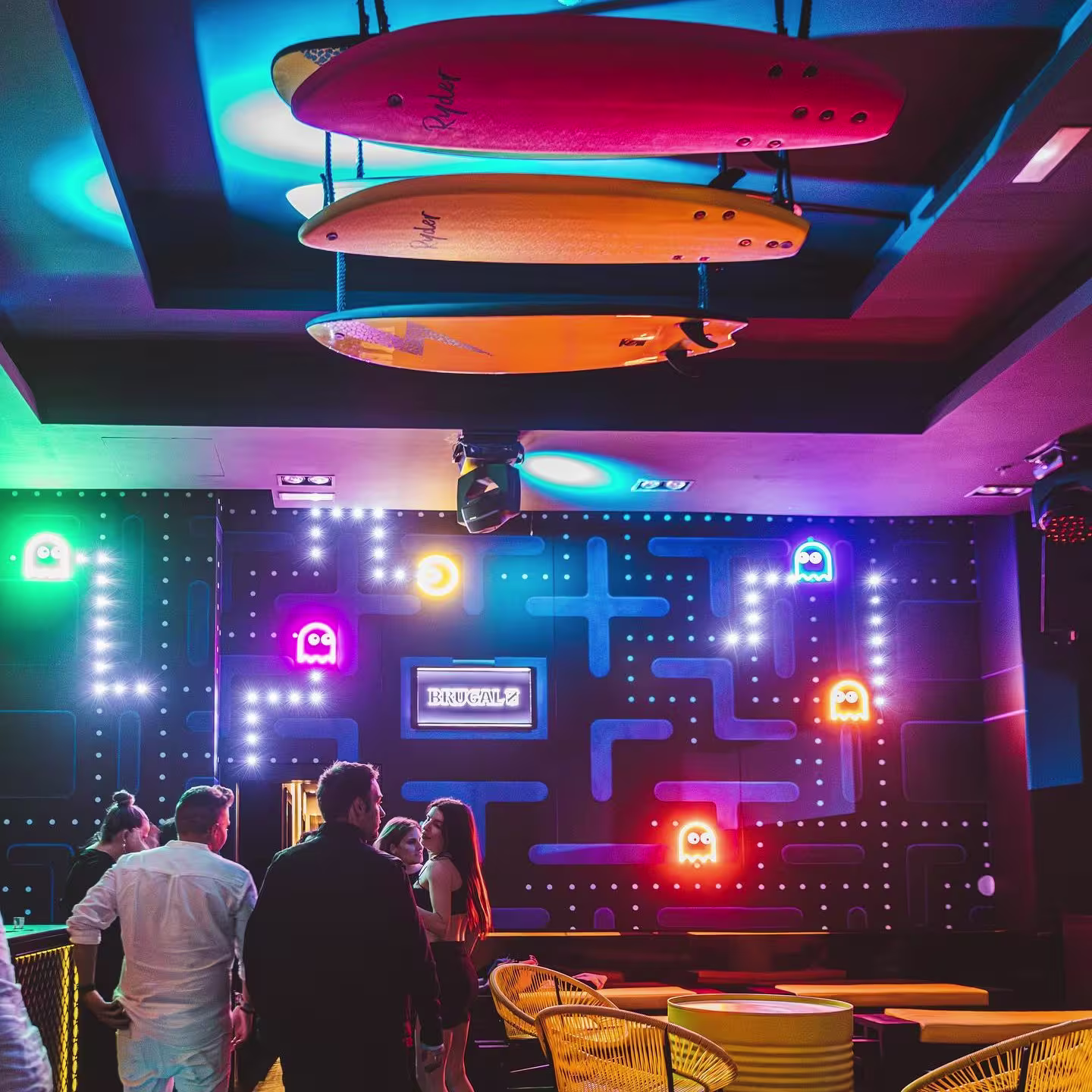 Hyp3: Bar de ambiente retro-futurista con paredes inspiradas en Pac-Man y tablas de surf en el techo. Los colores neón y la iluminación crean un espacio divertido y dinámico para los amantes del estilo arcade.