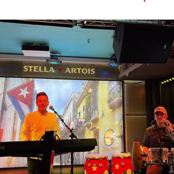 La imagen muestra un entorno interior con una configuración para una actuación musical en La Negra Tomasa. Hay una persona sentada frente a un teclado con un soporte de micrófono delante. se observa una candera grande de cuba en el fondo.