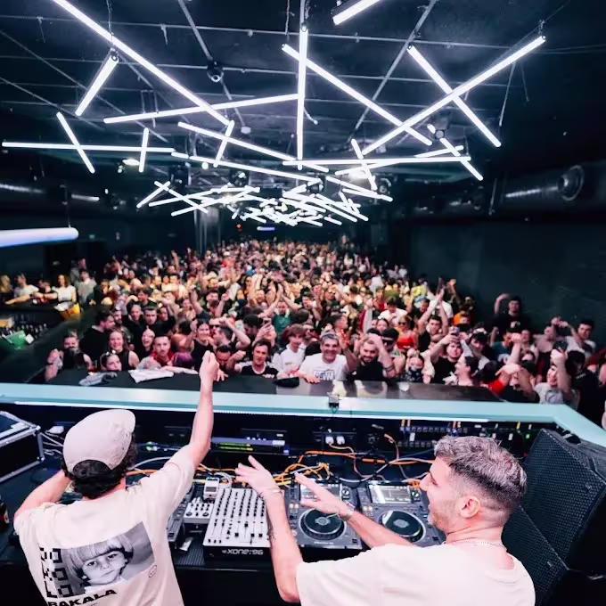 Cabina de DJ en Sala Sonora con dos pinchadiscos animando a una gran multitud, bajo techo oscuro iluminado con tiras de luz en forma geométrica.