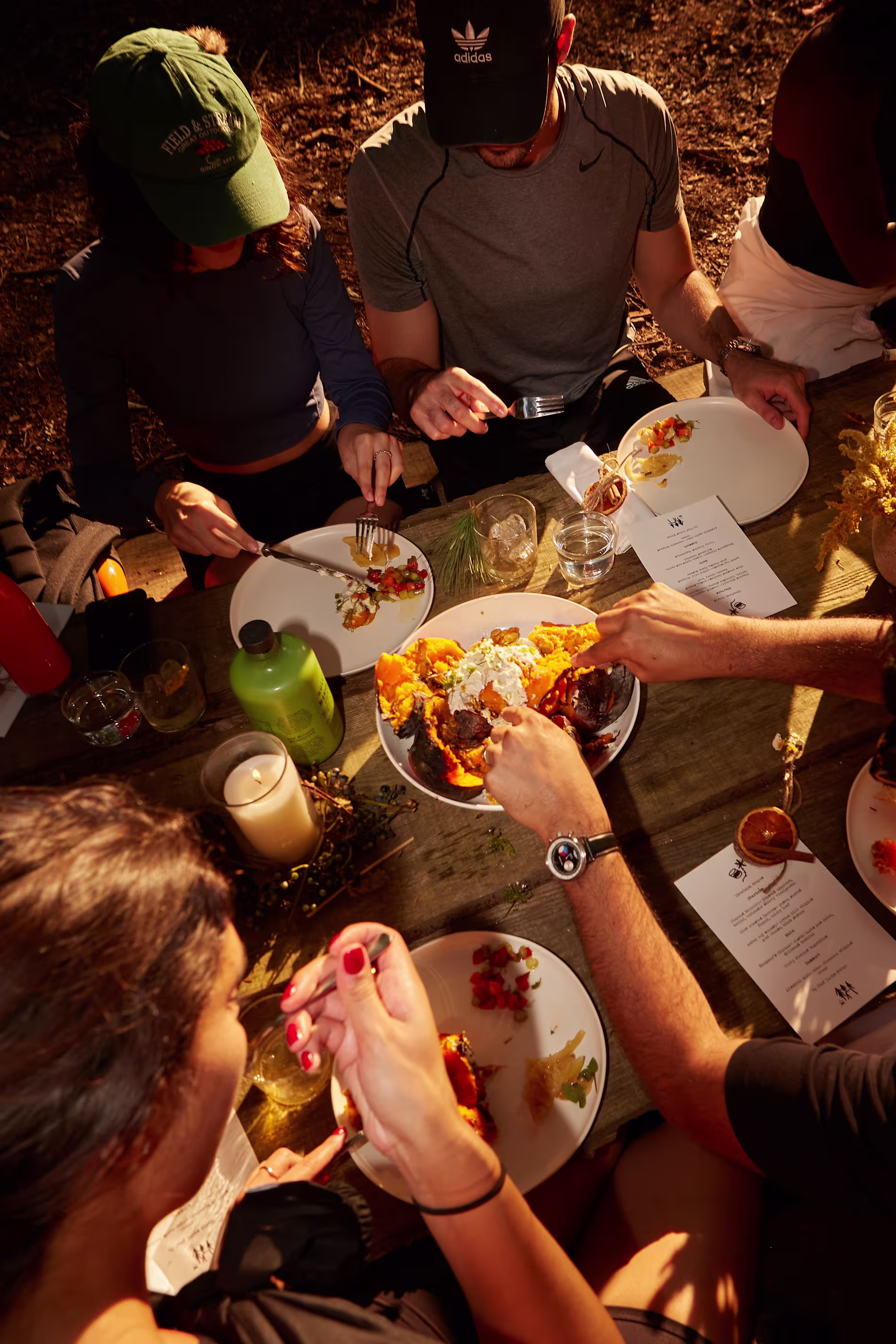 Four people sit at a wooden table outdoors, eating food from plates with utensils. Menus, drinks, and a candle are visible on the table in warm sunlight.