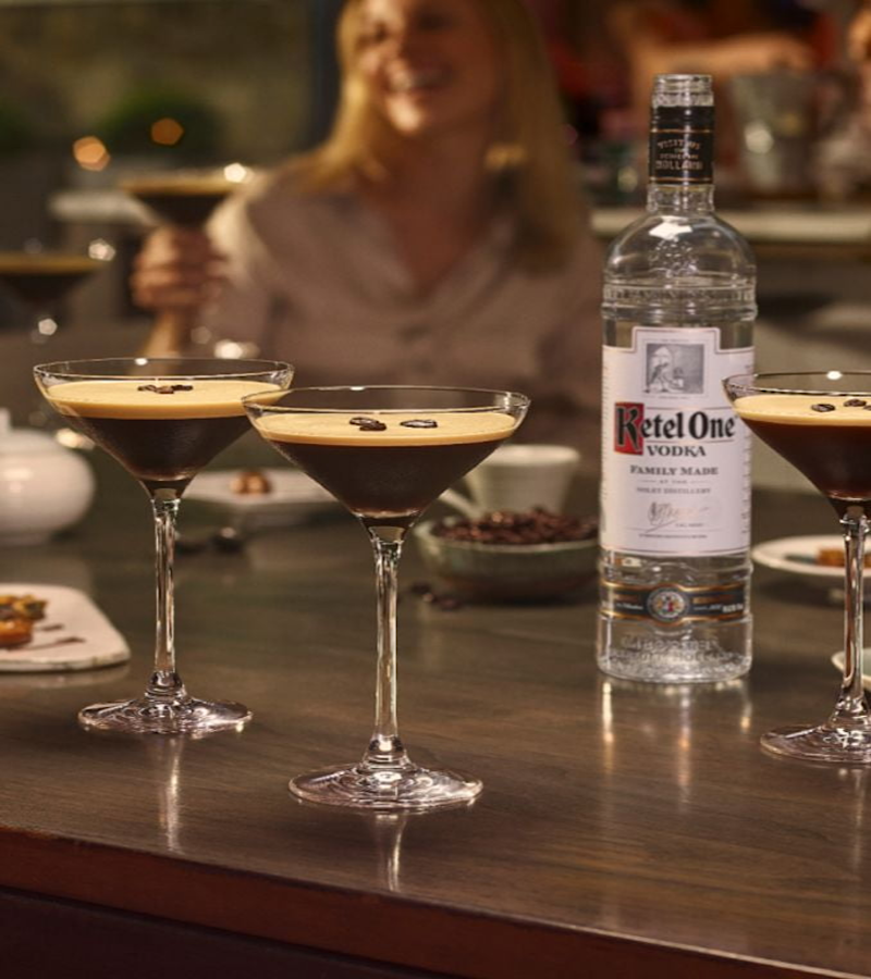 Espresso martini cocktails served in coupe glasses with coffee beans, next to a bottle of Ketel One Vodka on a bar table.