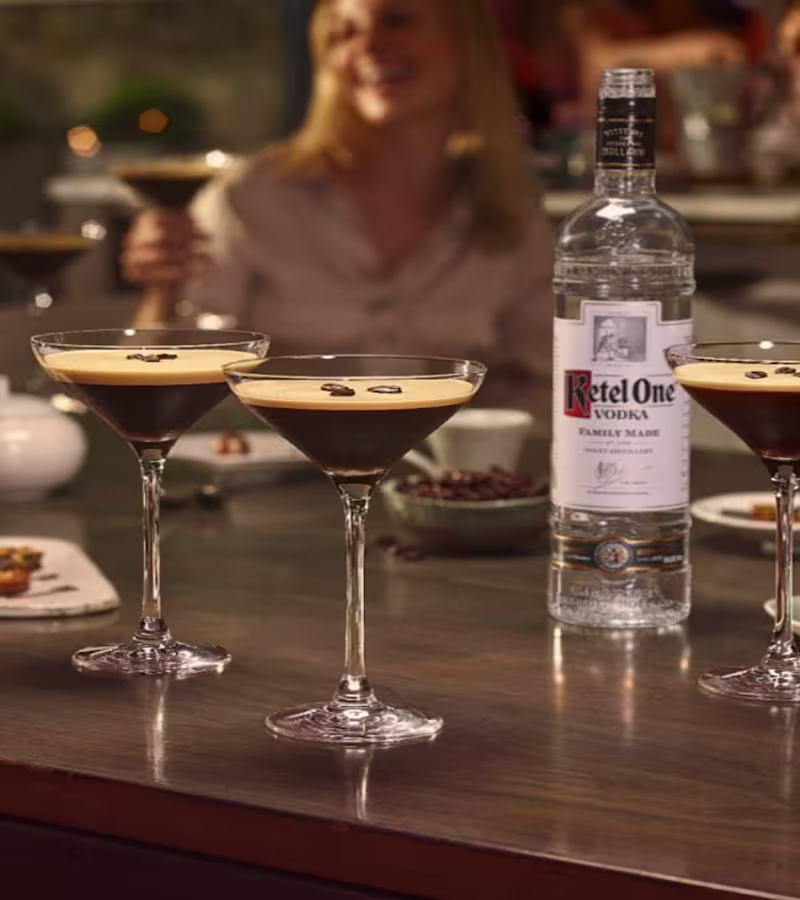 Espresso martini cocktails served in coupe glasses with coffee beans, next to a bottle of Ketel One Vodka on a bar table.