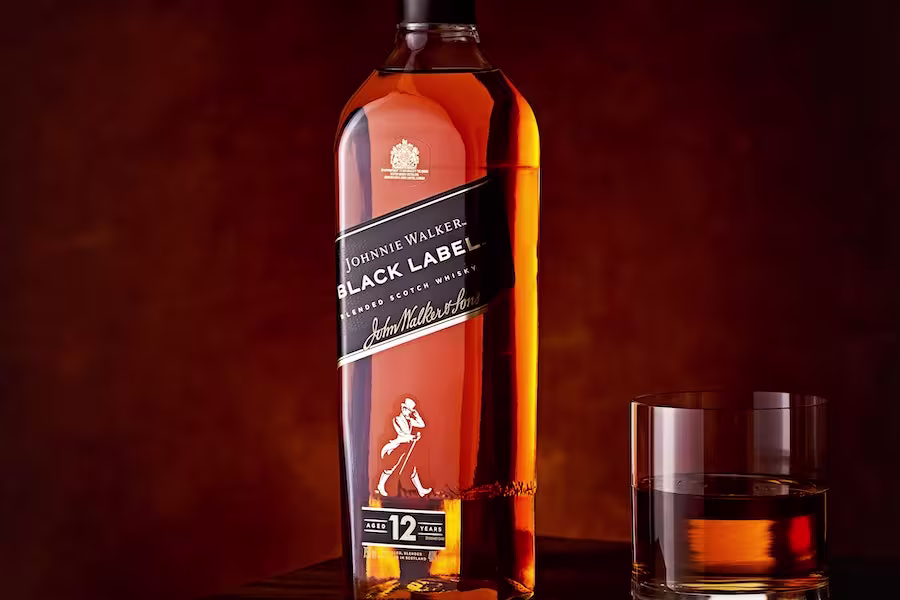 A bottle of Johnnie Walker Black Label 12-year-old blended Scotch whisky stands next to a glass with whisky, set against a dark brown background.