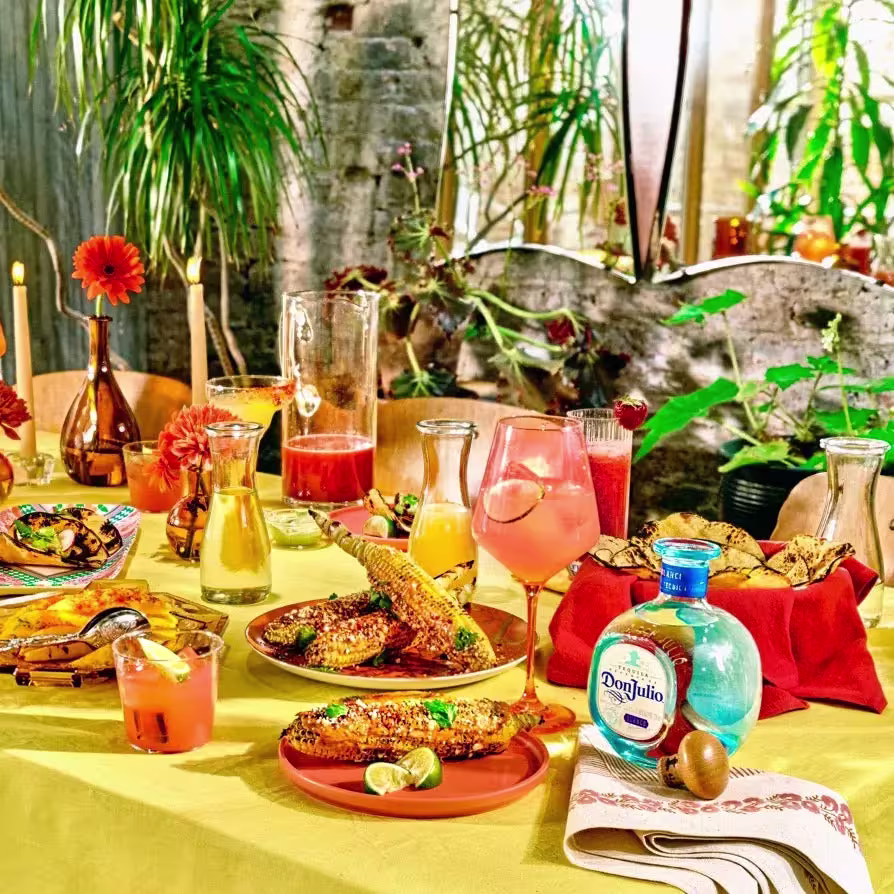 A festive table set with Mexican food, colorful drinks, grilled corn, candles, a bottle of Don Julio tequila, and vibrant decorations, surrounded by plants.