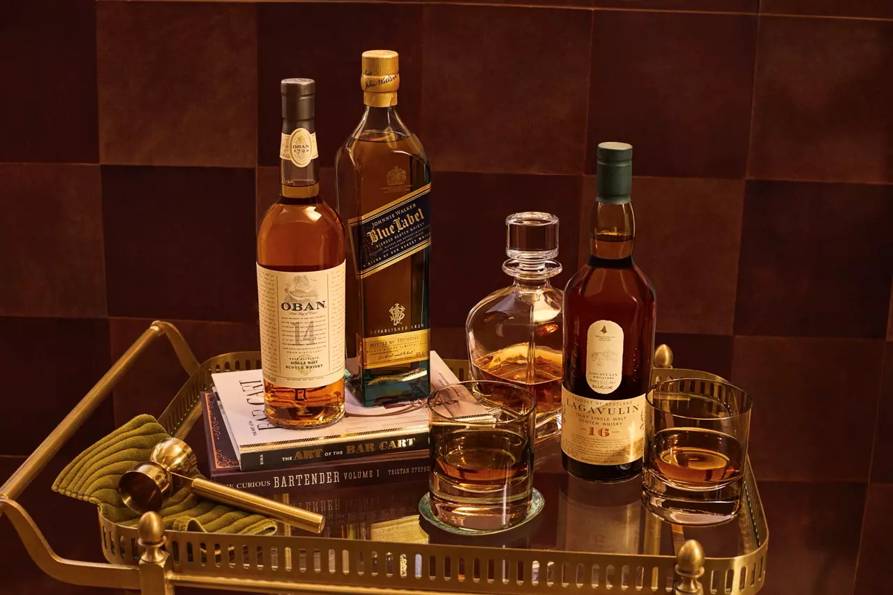 A brass tray holds bottles of Oban, Johnnie Walker Blue Label, and Lagavulin 16 whisky, a decanter, filled glasses, bar tools, and cocktail books on a brown surface.