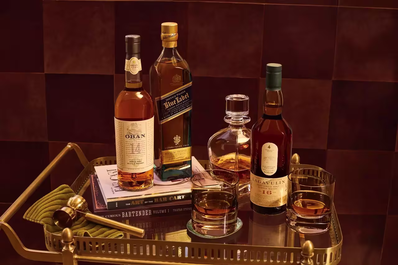 A brass tray holds bottles of Oban, Johnnie Walker Blue Label, and Lagavulin 16 whisky, a decanter, filled glasses, bar tools, and cocktail books on a brown surface.