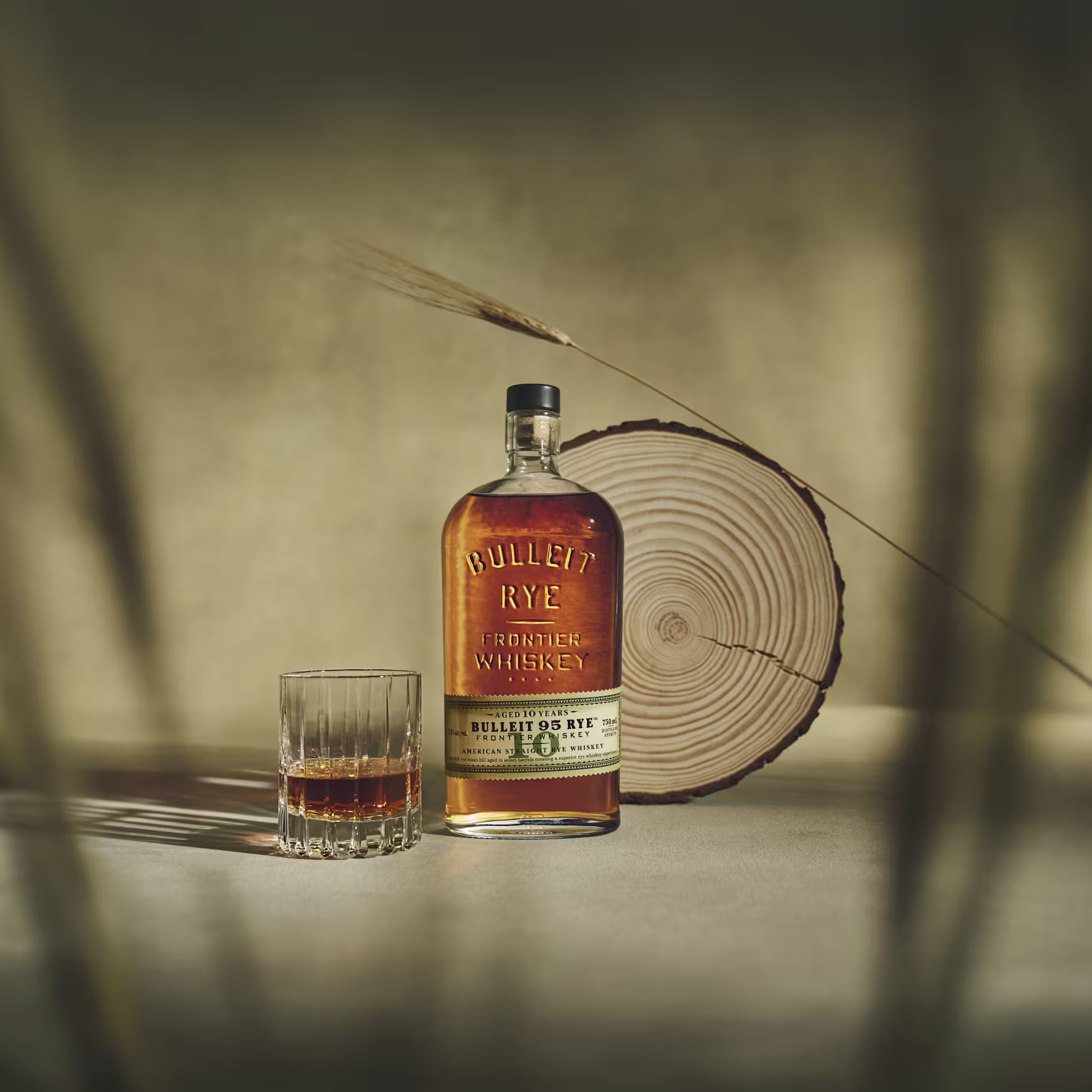 A bottle of Bulleit Rye Frontier Whiskey and a glass with whiskey sit on a surface beside a cross-section of a tree trunk, with soft shadows and a neutral background.