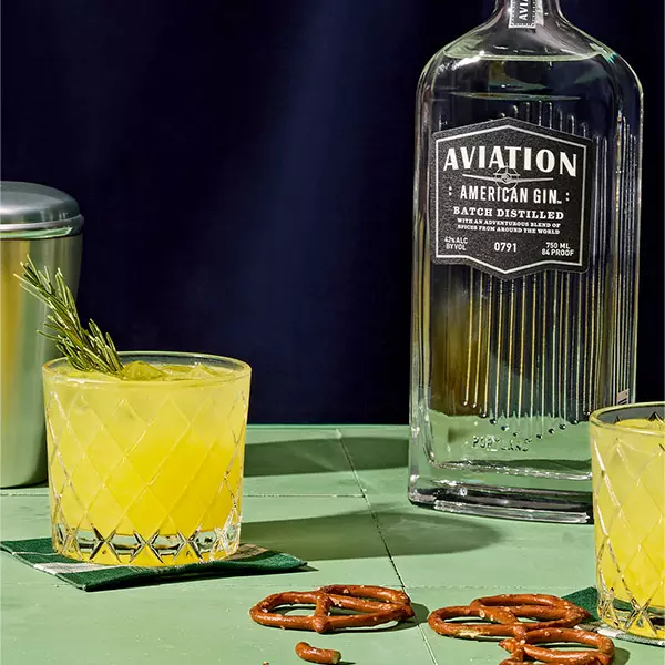 A glass of yellow cocktail garnished with rosemary sits on a green coaster beside a bottle of Aviation American Gin, pretzels, and another glass on a green table.