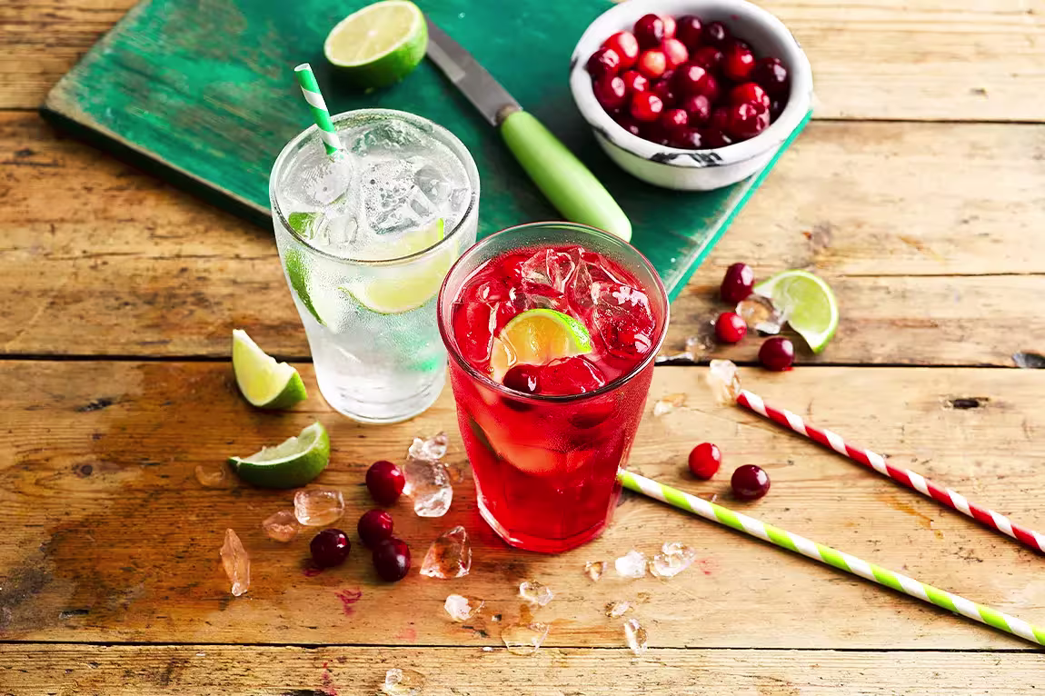 Two glasses of drinks with ice and lime wedges on a wooden table, one clear and one red. Scattered cranberries, lime slices, a bowl of cranberries, and striped straws nearby.