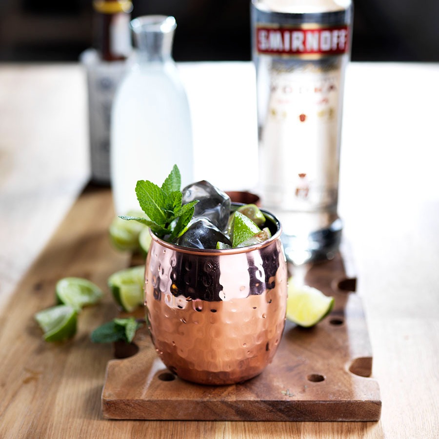 A Moscow Mule cocktail in a copper cup on a wooden cutting board, next to a bottle of Smirnoff vodka.