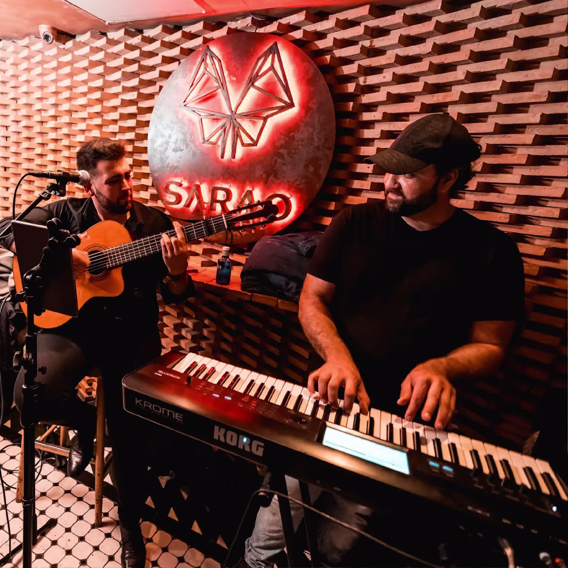 La imagen muestra a dos músicos en vivo en Sarao, uno tocando la guitarra y el otro el teclado, con el logo iluminado de "Sarao" en el fondo.