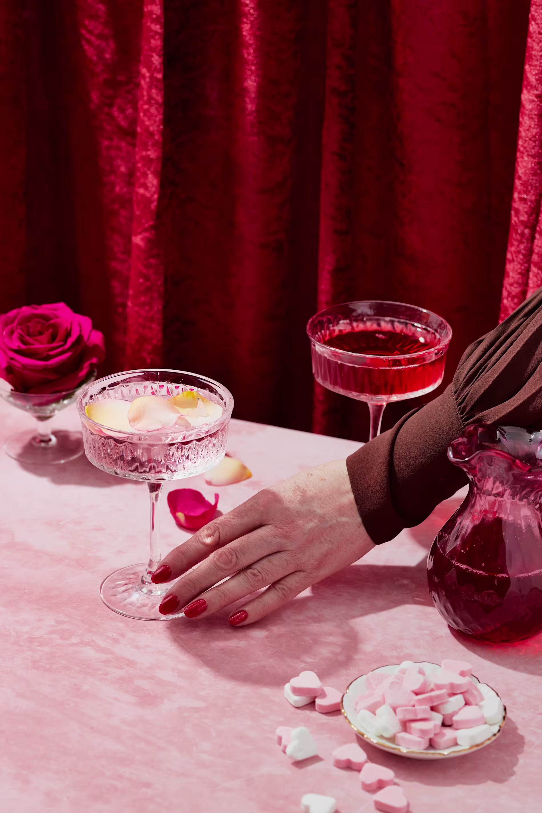 A hand with red nails rests on a pink table with two cocktails, a red rose, a glass pitcher, and heart-shaped candies, set against a red velvet curtain.