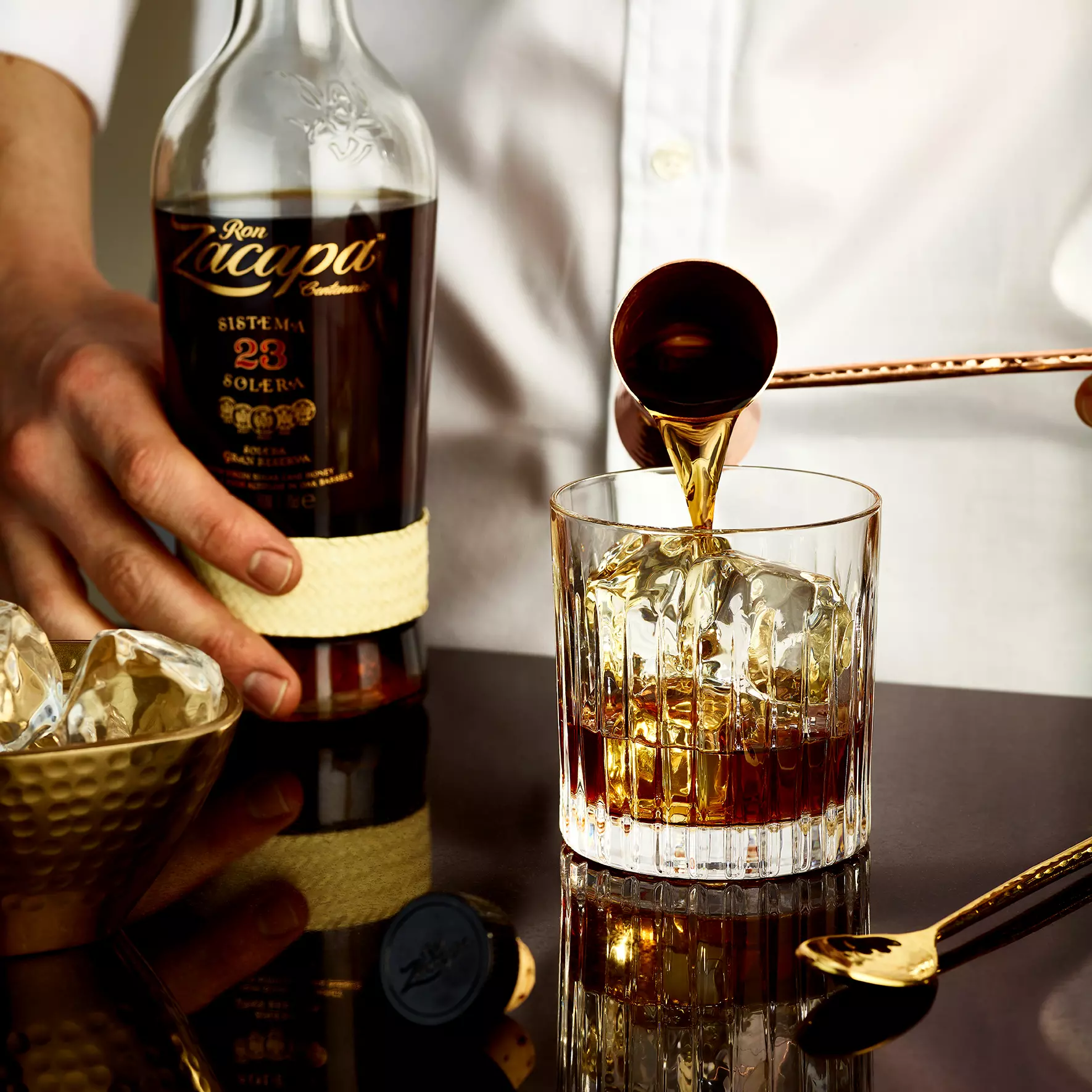 A person pours Ron Zacapa rum from a bottle into a glass with ice cubes, with a golden spoon and bowl nearby on a reflective surface.