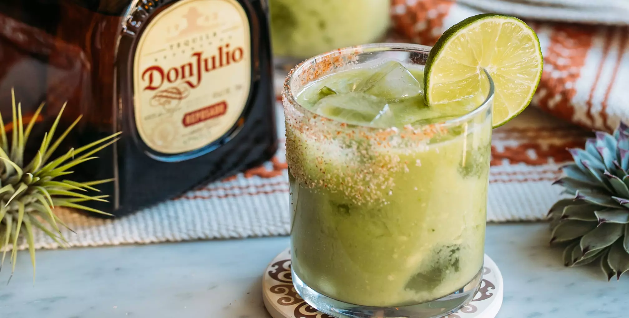 A glass of green cocktail with ice and a lime wheel garnish is on a coaster, next to a bottle of Don Julio tequila.