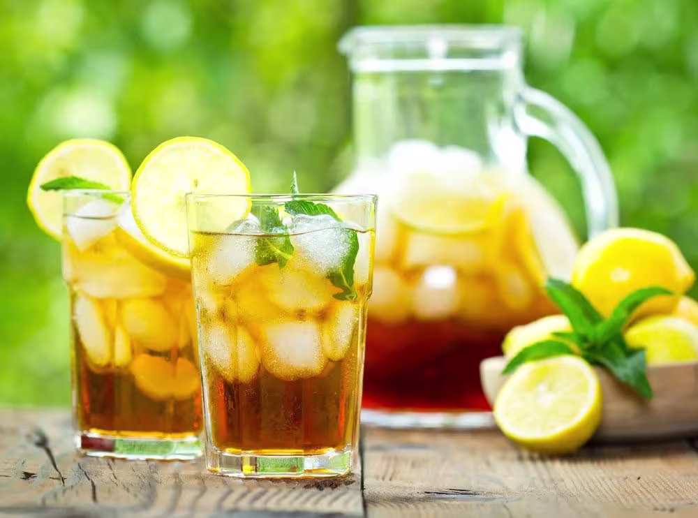 Two glasses of iced tea with lemon slices, mint leaves, and ice sit on a wooden table next to a pitcher of iced tea and a bowl of lemons.
