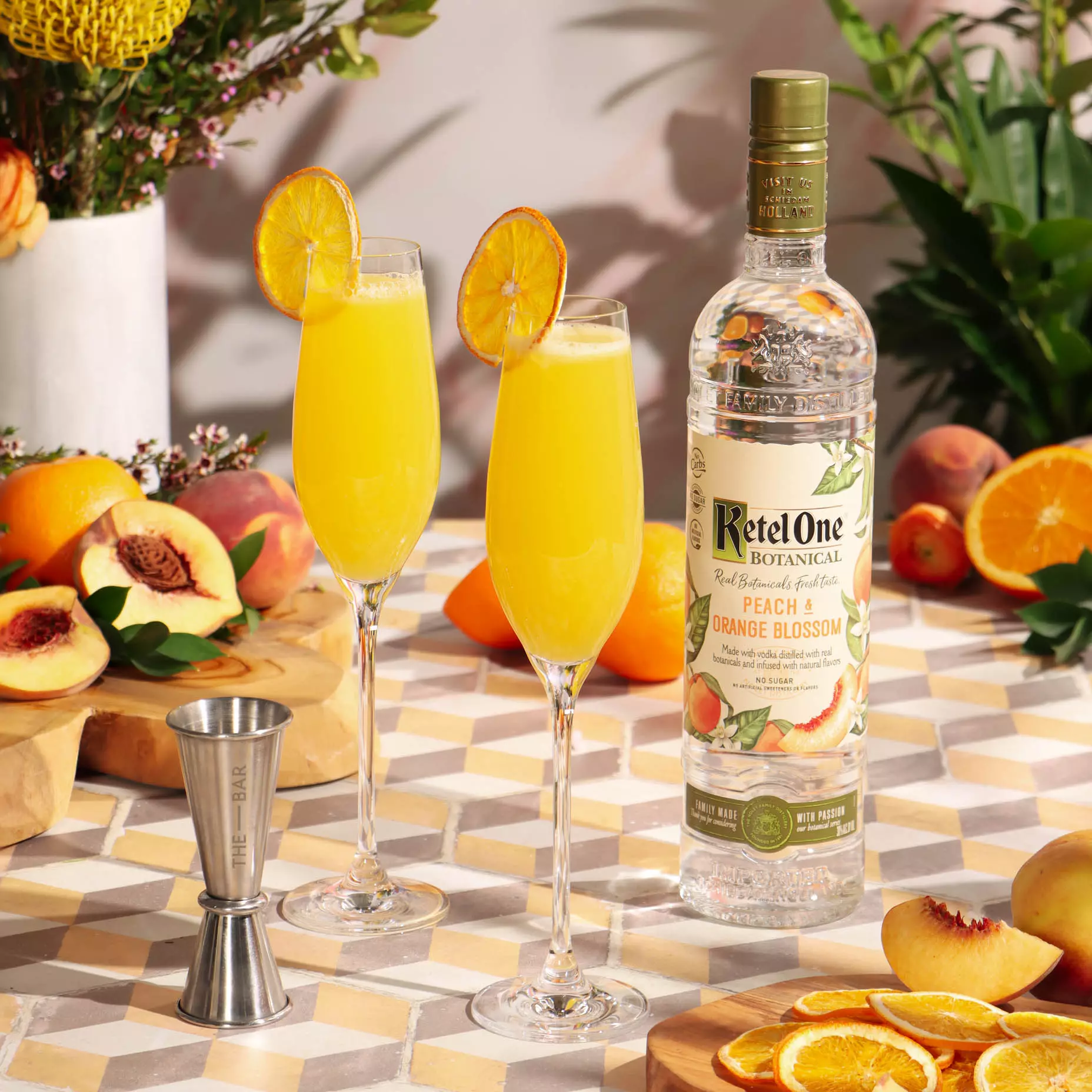 Two glasses of orange cocktail garnished with dried orange slices beside a bottle of Ketel One Botanical Peach & Orange Blossom vodka, surrounded by fresh peaches and oranges on a tiled surface.