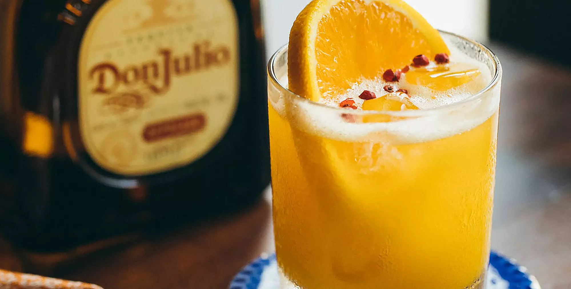 A glass of orange cocktail garnished with an orange slice and red peppercorns sits next to a bottle of Don Julio tequila.